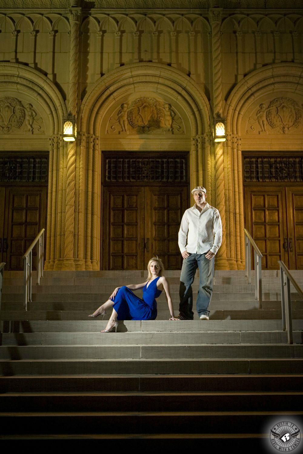 hot blond with long blue dress and tan heels sits seductively on steps near a standing guy with a cab driver hat wearing a light blue button up shirt and blue jeans also on steps in front of a gothic church with arched stone work and several large wooden doors in this intense Houston engagement picture